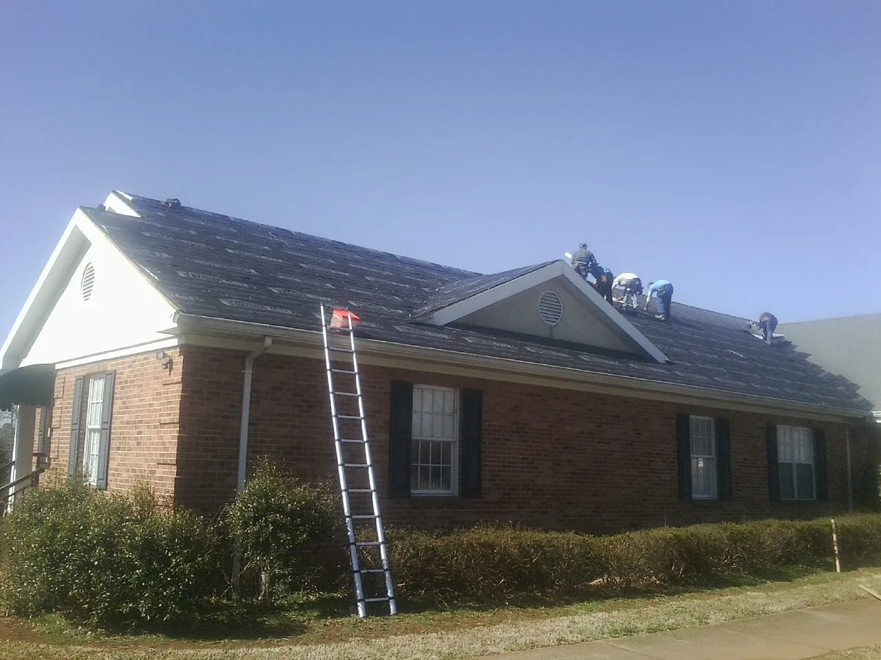 Crew tearing shingles off a brick ranch roof with a ladder set — Metal Roof Repair work by our crew in East Amherst