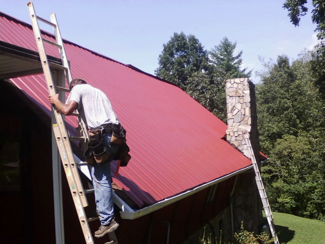 Roofer on a ladder beside a red metal roof and stone chimney — Soffit & Fascia Repair by Roofing Brothers in East Amherst